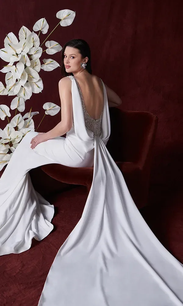 Eine Braut sitzt in einem rückenfreien Hochzeitskleid mit Perlenverzierung auf einem roten Sofa. Der Hintergrund ist tiefrot, dekoriert mit weißen Blumen.