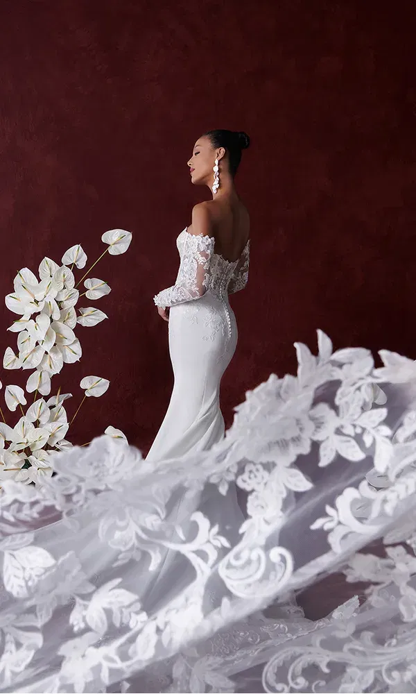 Eine Braut in einem schulterfreien Spitzenhochzeitskleid, steht seitlich vor einem tiefroten Hintergrund. Der Rock ist dramatisch in Bewegung, umgeben von weißen Blumen.