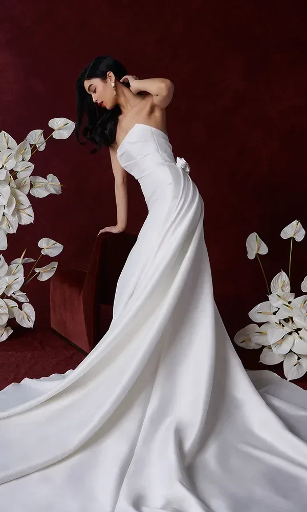 Eine Braut in einem eleganten, schulterfreien Hochzeitskleid mit langer Schleppe, lehnt an einem roten Sofa. Der Hintergrund ist tiefrot und mit weißen Blumen dekoriert.
