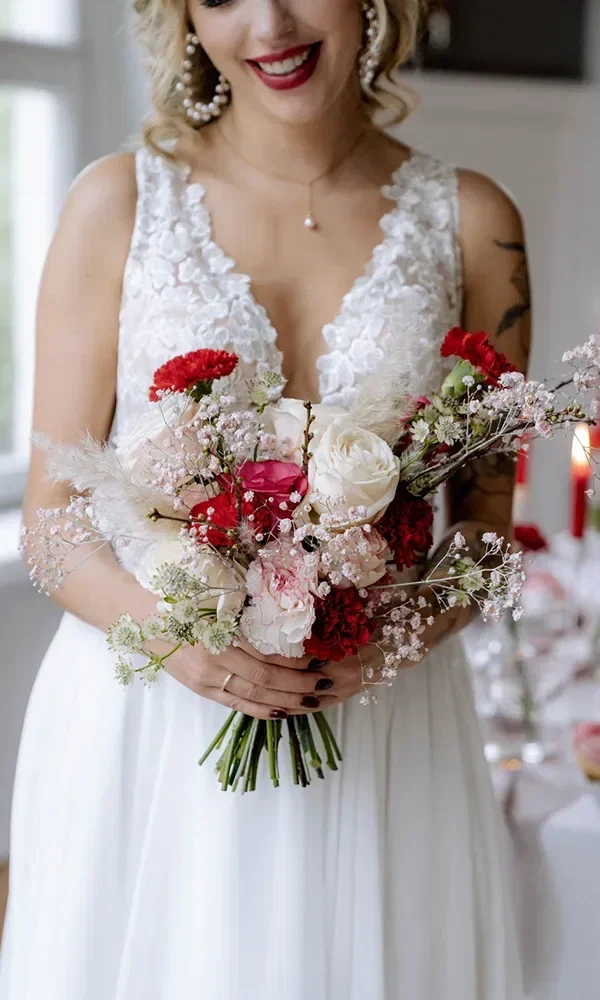 Eine Braut hält einen wunderschönen Strauß aus roten und weißen Blumen, mit zarten rosa Akzenten und grünen Blättern.