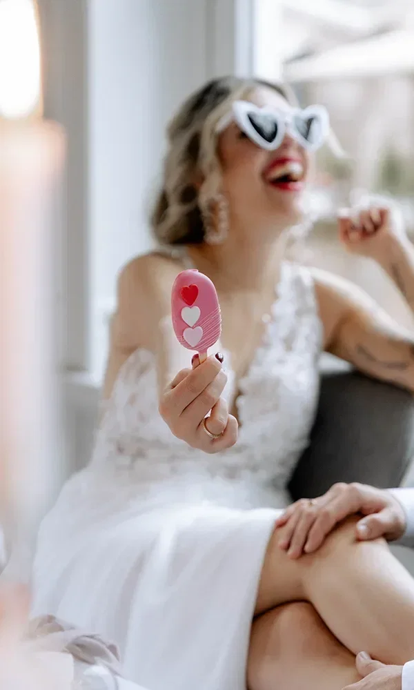 Eine Braut hält einen rosa Cakepop mit Herzverzierungen und trägt dabei herzförmige Sonnenbrillen.