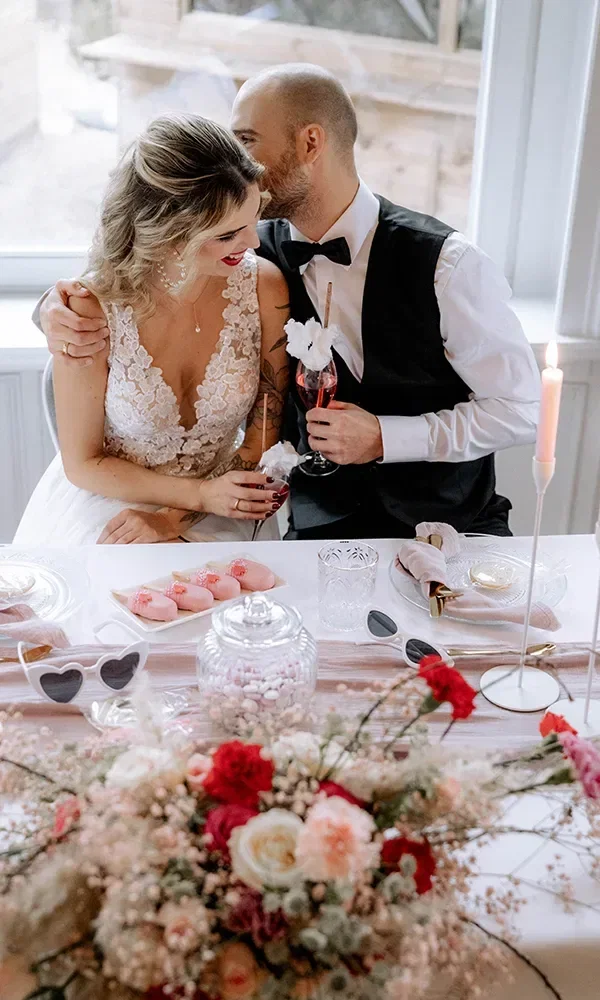 Ein Brautpaar sitzt an einem elegant gedeckten Tisch. Die Braut trägt ein wunderschönes weißes Spitzenkleid und lächelt, während der Bräutigam, gekleidet in einem schwarzen Anzug mit Fliege, ihr zärtlich etwas ins Ohr flüstert. Vor ihnen auf dem Tisch stehen rosa glasierte Donuts, elegante Gläser, Herz-Sonnenbrillen und eine schöne Blumendekoration in Rosa und Rot.