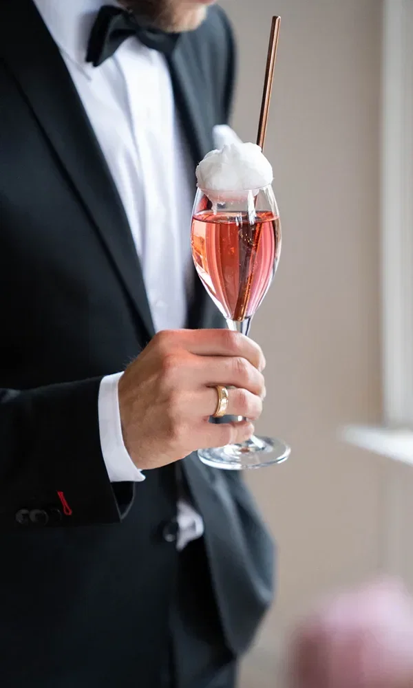 Ein Mann im schwarzen Anzug hält ein Glas Sekt mit einer weißen Zuckerwattegarnitur.