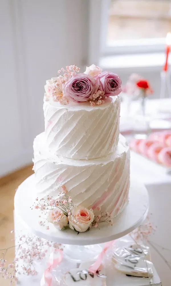 Eine zweistufige Hochzeitstorte, dekoriert mit rosa und lila Blumen, auf einem weiß gedeckten Tisch.
