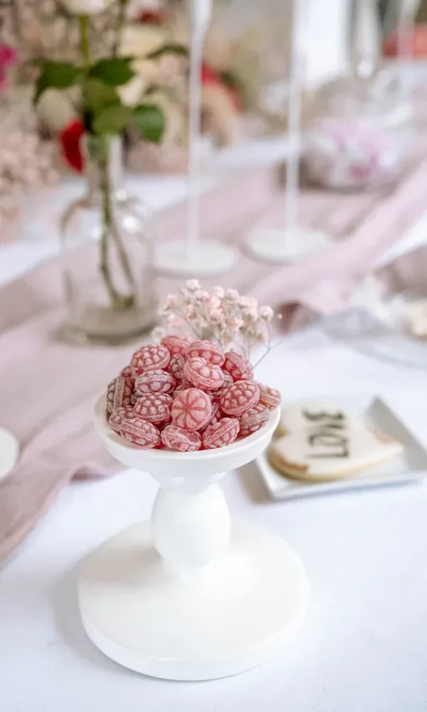Eine Schale voller rosafarbener Bonbons, dekoriert mit zarten rosa Blüten auf einem elegant gedeckten Tisch.