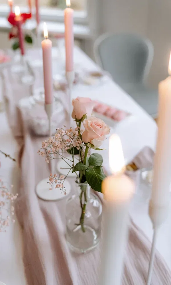 Ein elegant gedeckter Tisch mit brennenden rosa Kerzen und zarten Rosen in einer Glasvase, drapiert mit einem sanft rosa Tischtuch.
