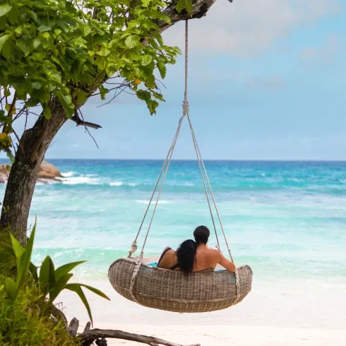 Ein Paar sitzt in einer Hängeschaukel am Strand mit Blick auf das türkisblaue Meer.