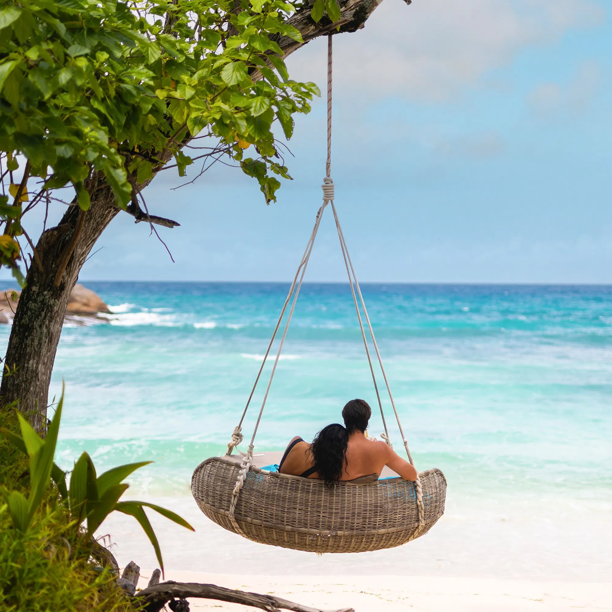 Ein Paar sitzt in einer Hängeschaukel am Strand mit Blick auf das türkisblaue Meer.