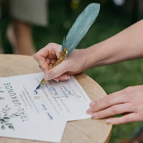 Eine Nahaufnahme der Braut, die mit einem dekorativen Federkiel eine Heiratsurkunde unterzeichnet. Die Urkunde ist mit floralen Mustern verziert und hat das Wort 'TRAU-URKUNDE' deutlich sichtbar. Die Hand der Braut und ein Teil ihres blauen Kleides sind zu sehen, im Hintergrund befindet sich eine grüne Pflanze.