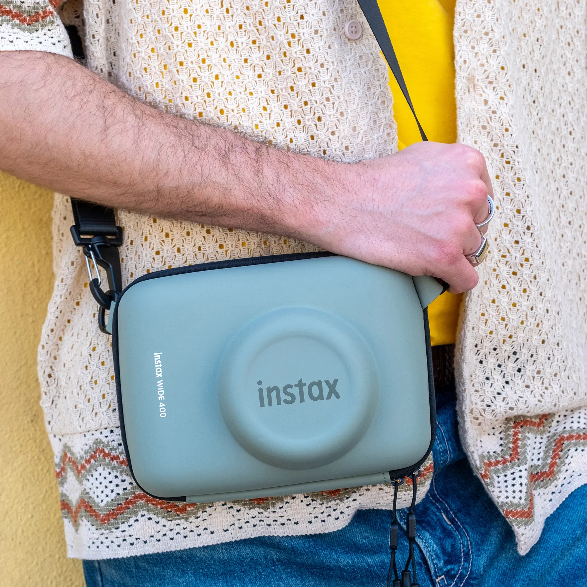 Eine Person trägt eine Instax Wide 400 Kamera in einer grünen Tragetasche über der Schulter. Die Tasche ist aus robustem Material und hat einen breiten Schultergurt. Der Fokus liegt auf der Tasche, während der Oberkörper der Person in einem lässigen, gestrickten Oberteil mit einem gelben T-Shirt darunter zu sehen ist. Die Hand der Person hält die Tasche fest an der Seite.