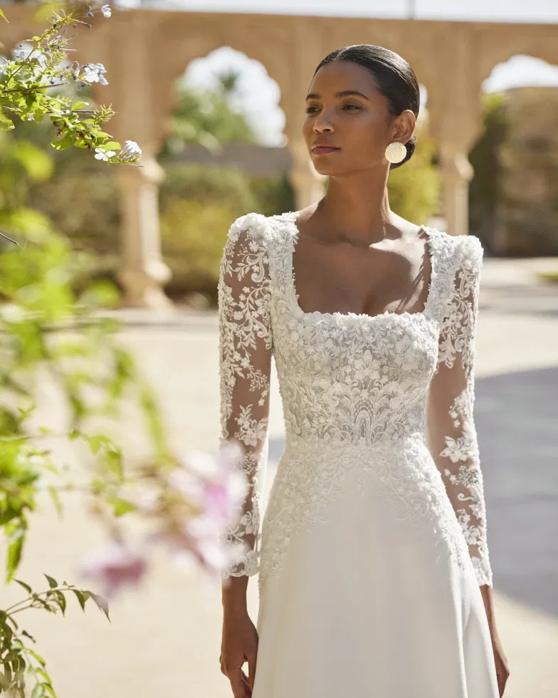 Elegante Braut in einem figurbetonten Brautkleid mit floraler Spitze, langen Ärmeln und eckigem Ausschnitt, fotografiert in einer sonnigen Arkadenkulisse mit mediterranem Flair.