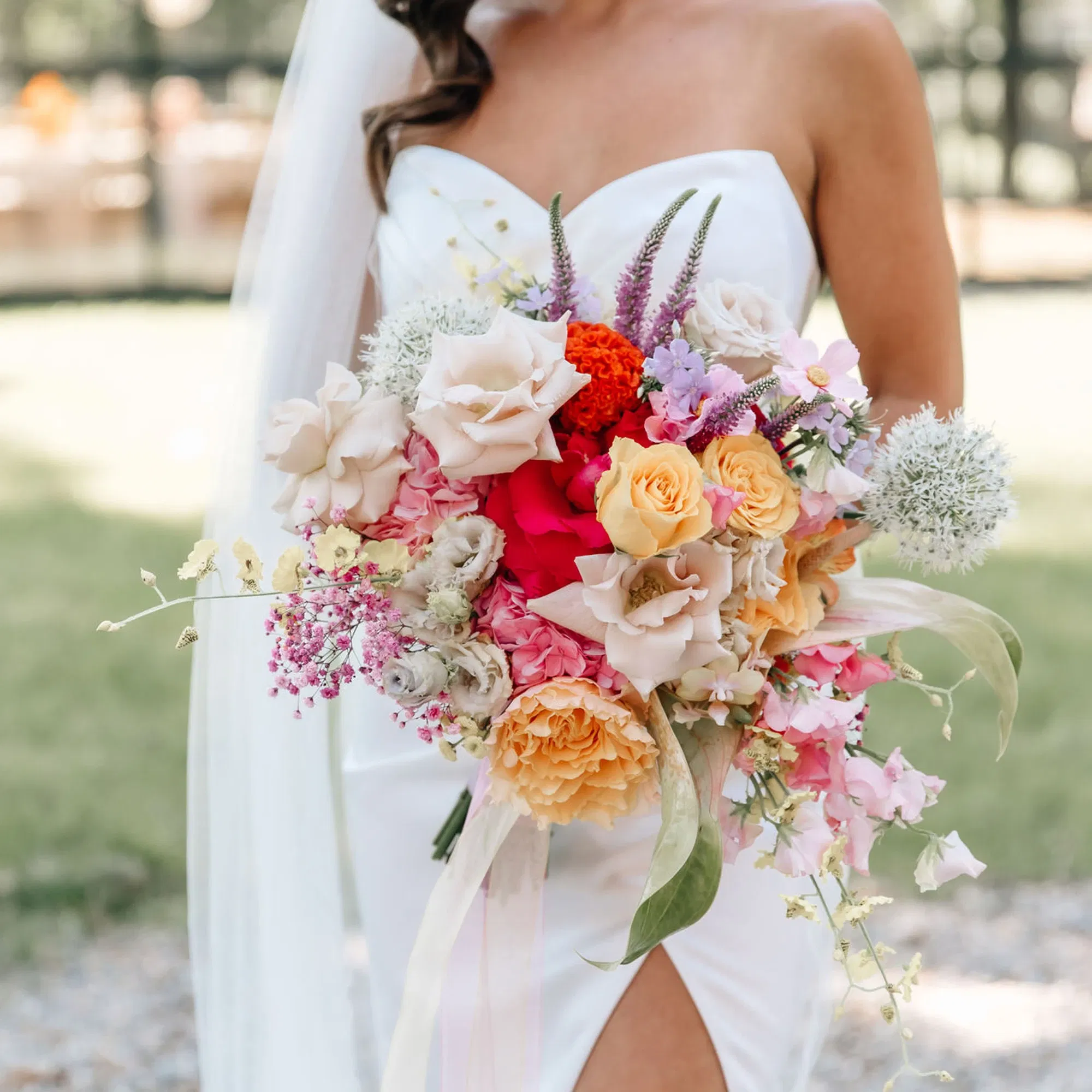 Eine Braut in einem trägerlosen weißen Brautkleid hält einen großen, bunten Blumenstrauß. Der Strauß enthält Rosen in Gelb, Rosa und Creme, ergänzt durch lilafarbene und weiße Blüten. Im Hintergrund sieht man einen grünen Garten mit unscharfen Details.