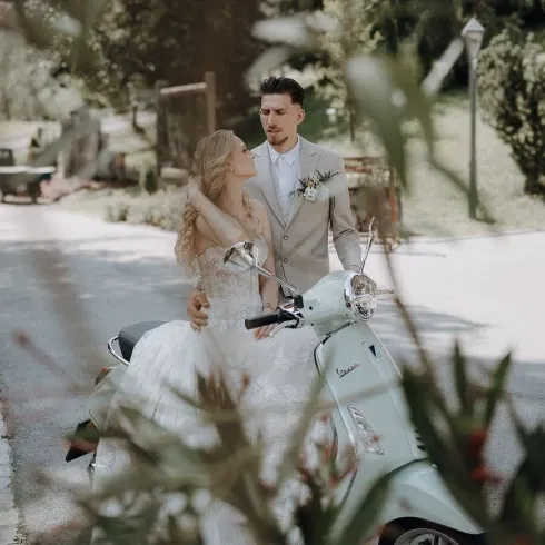 Braut und Bräutigam stehen romantisch an einer Vespa, die Braut sitzt auf dem Roller, während der Bräutigam sie liebevoll umarmt.