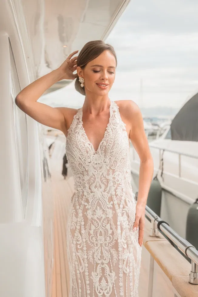 Elegante Braut in einem figurbetonten Brautkleid mit tiefem V-Ausschnitt und opulenter Spitze, die entspannt an der Reling einer Yacht steht und von sanftem Tageslicht umgeben ist.