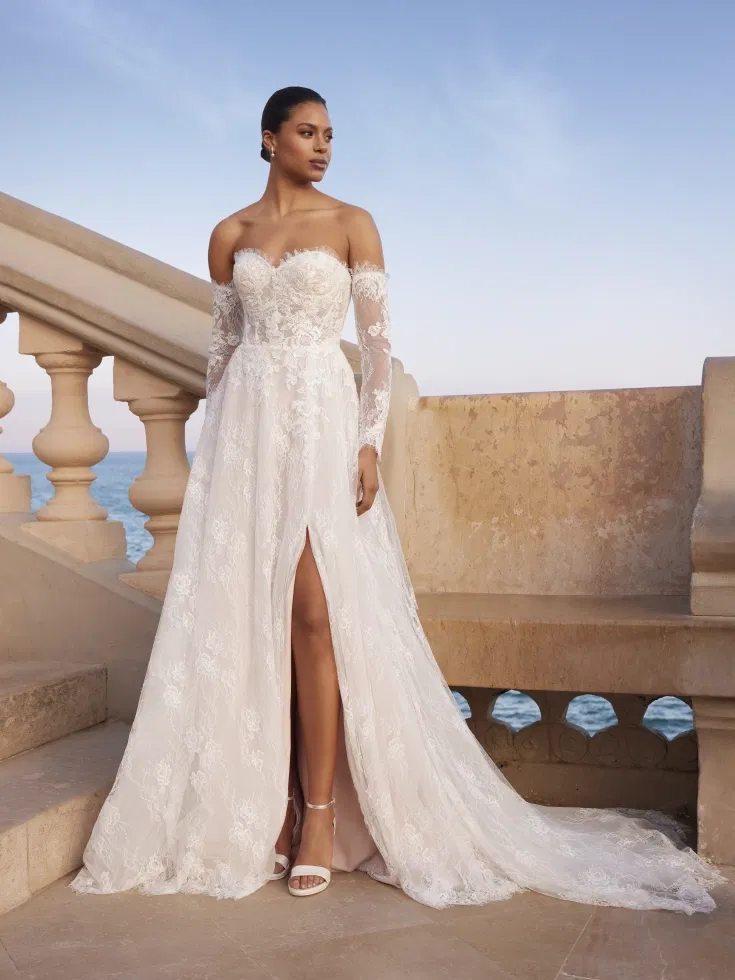 Elegante Braut in einem schulterfreien Brautkleid mit langen Spitzenärmeln, Herzausschnitt und hohem Beinschlitz, inszeniert auf einer mediterranen Terrasse mit Meerblick und warmem Abendlicht.