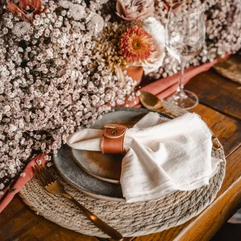 Eleganter Hochzeitstisch mit natürlichen Materialien: Leinenserviette in einem Lederserviettenring, platziert auf einem handgefertigten Teller und umgeben von Blumenschmuck aus Schleierkraut.