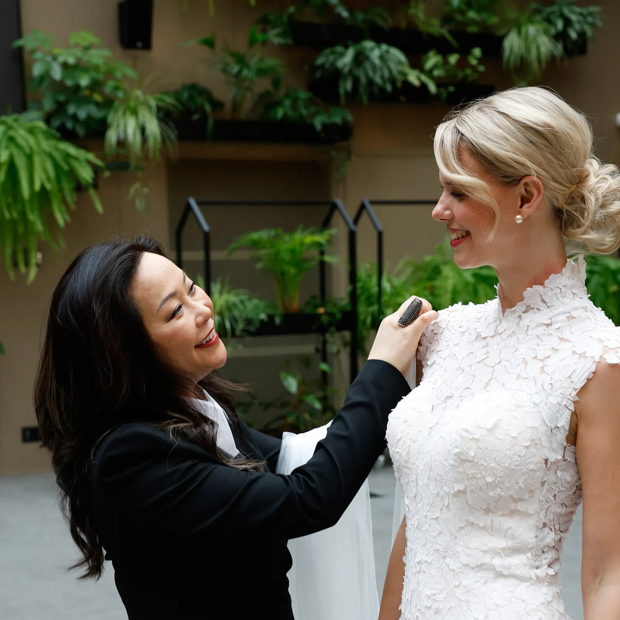 Die Brautmodendesignerin Sabrina Toy passt das Kleid einer lächelnden Braut an. Die Braut trägt ein elegantes weißes Spitzenkleid, während die Designerin in Schwarz gekleidet ist. Im Hintergrund sind grüne Pflanzen zu sehen, die der Szene eine frische und natürliche Atmosphäre verleihen.