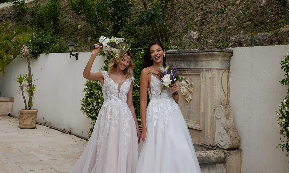 Zwei Bräute, die fröhlich in romantischen Spitzenkleidern stehen, eine davon mit hochgehaltenem Brautstrauß. Beide stehen vor einem dekorativen Steinbrunnen.