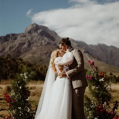 Lina Mallon und Chris bei ihrer Trauung auf einer Wiese in Kapstadt, umgeben von Blumen und Weinbergen, mit einer malerischen Bergkulisse im Hintergrund.