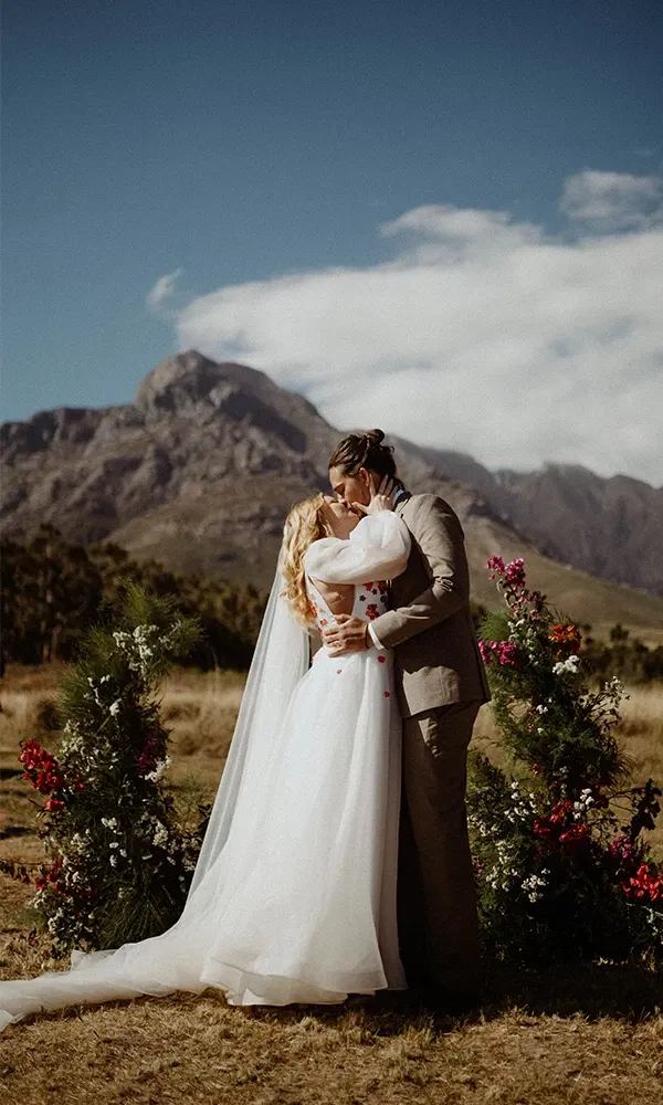 Lina Mallon und Chris bei ihrer Trauung auf einer Wiese in Kapstadt, umgeben von Blumen und Weinbergen, mit einer malerischen Bergkulisse im Hintergrund.