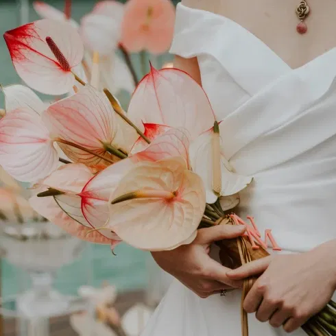 Braut mit schlichtem weißen Kleid hält einen Brautstrauß aus zarten, pastellfarbenen Anthurien-Blüten mit einem Hauch von Rot, fotografiert in einer eleganten, modernen Hochzeitsumgebung.