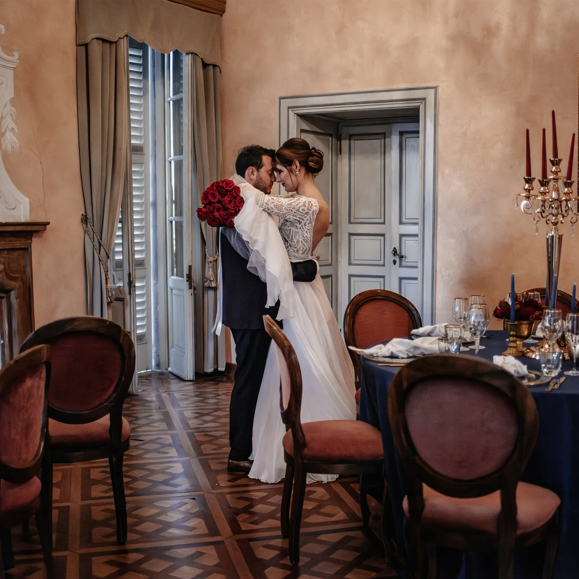 Braut und Bräutigam umarmen sich in einem eleganten Raum mit antiken Möbeln und einem runden Tisch, der mit roten und blauen Details dekoriert ist.