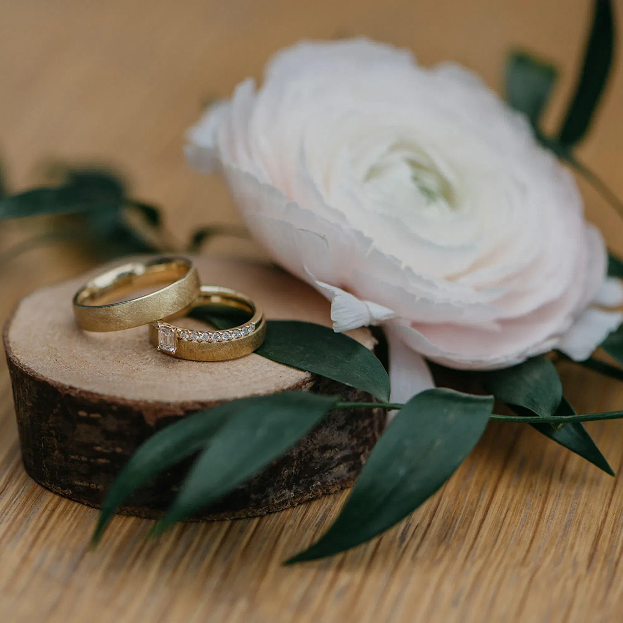 Goldene Eheringe auf einer Holzscheibe, dekoriert mit einer weißen Blume und grünen Blättern, perfekt für eine stilvolle Hochzeitsdekoration.