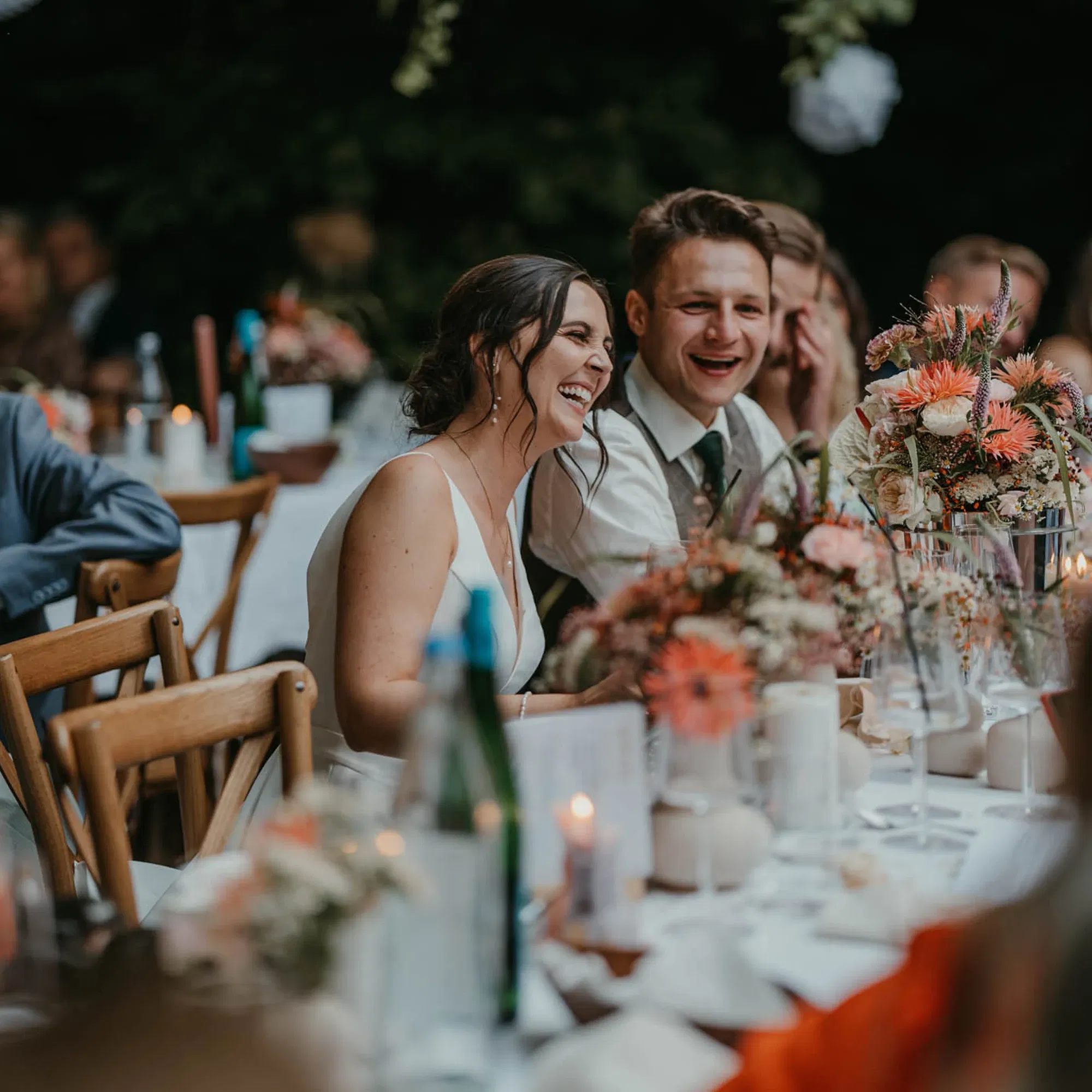 Ein Brautpaar sitzt bei ihrer Hochzeitsfeier an einem festlich gedeckten Tisch im Freien und lacht herzhaft. Der Tisch ist mit Blumenarrangements und Kerzen dekoriert, während die Gäste im Hintergrund entspannt und fröhlich wirken.