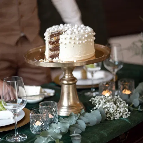 Weiße Hochzeitstorte mit Perlenverzierung, angeschnitten und präsentiert auf einem goldenen Tortenständer, umgeben von Kerzen und eleganter Tischdekoration.