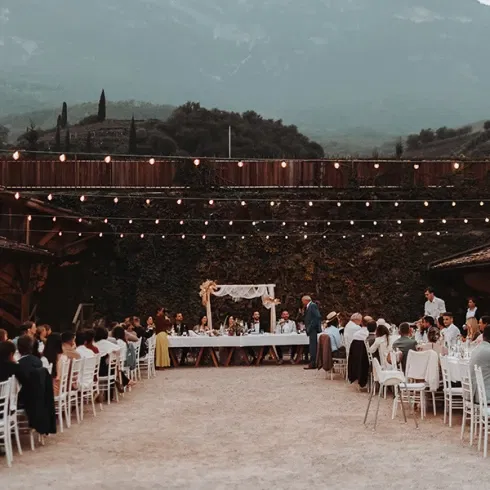 Hochzeitsempfang im Freien mit Tischen in U-Form, dekoriert mit weißen Tischdecken und Stühlen. Beleuchtet von Lichterketten, vor einer malerischen Bergkulisse, die eine entspannte, festliche Atmosphäre verleiht.