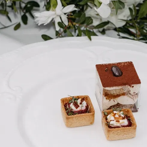 Fein angerichteter Dessertteller mit Tiramisu und zwei Mini-Tartelettes vor einem Blumenarrangement.