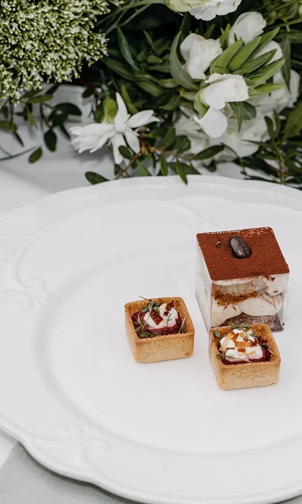 Fein angerichteter Dessertteller mit Tiramisu und zwei Mini-Tartelettes vor einem Blumenarrangement.