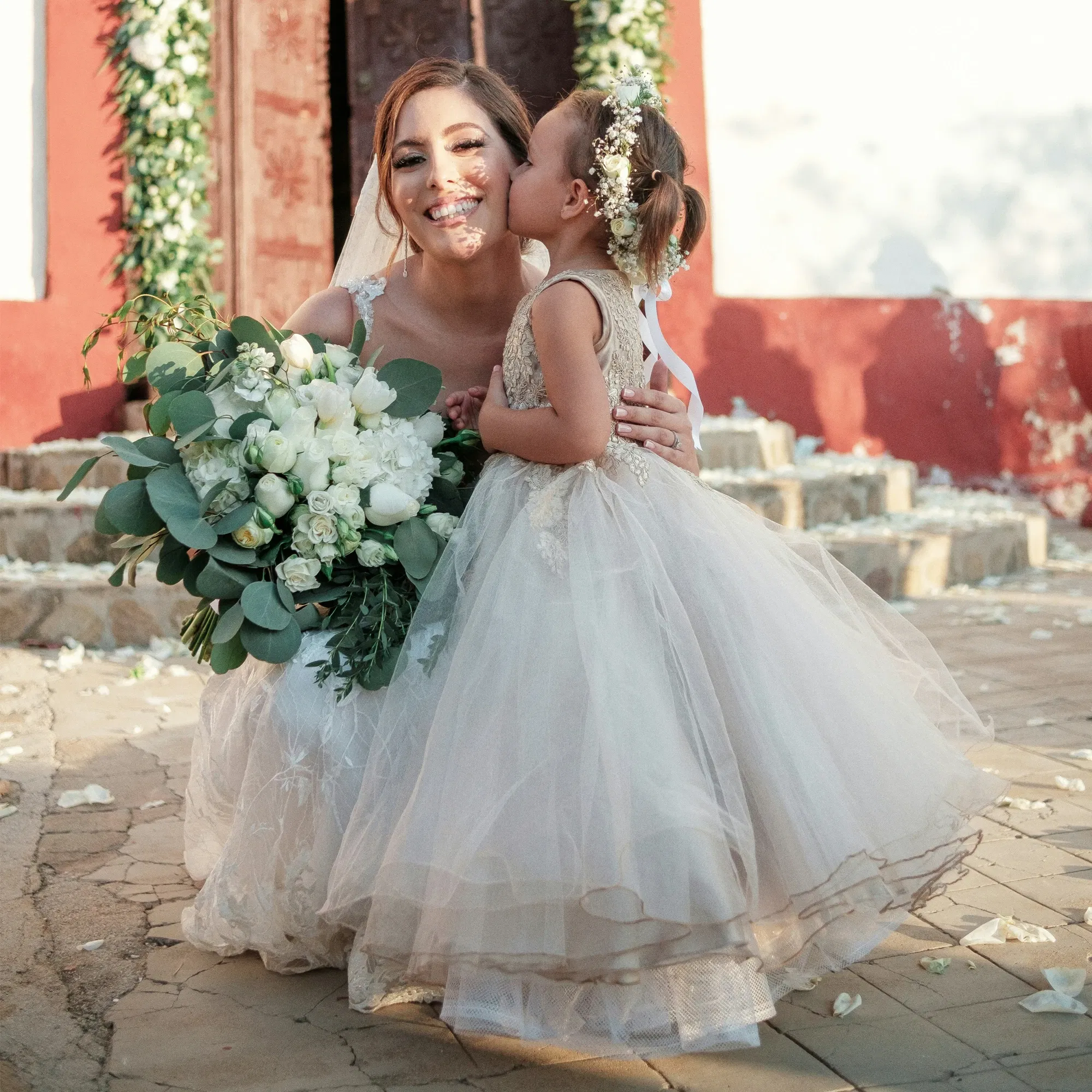 Braut, die lächelnd mit einem großen weißen Blumenstrauß kniet, während ein Blumenmädchen in einem Tüllkleid sie liebevoll auf die Wange küsst.