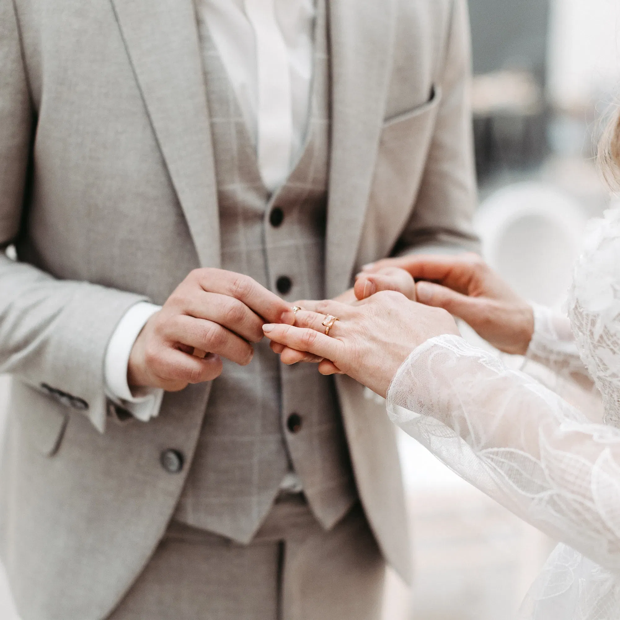 Ein Bräutigam im grauen Anzug steckt der Braut während der Hochzeitszeremonie den Ehering an den Finger. Die Braut trägt ein weißes Spitzenkleid, und die Szene zeigt den emotionalen Moment des Ringtauschs.