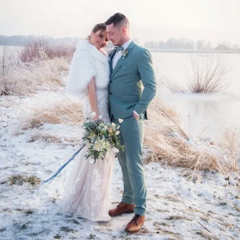 Ein Brautpaar posiert in einer verschneiten Landschaft am Ufer eines gefrorenen Sees. Die Braut trägt ein langes Spitzenkleid und eine weiße Stola, während der Bräutigam einen hellblauen Anzug trägt. Sie halten sich nah, und die Braut lächelt glücklich, während sie einen eleganten Blumenstrauß mit weißen Rosen und Eukalyptus hält.