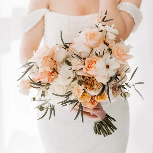Eine Braut in einem schulterfreien, perlweißen Kleid hält einen romantischen Brautstrauß mit pfirsichfarbenen und cremeweißen Rosen sowie grünen Zweigen. Der Strauß ist locker gebunden und steht im Kontrast zum eleganten Kleid der Braut.