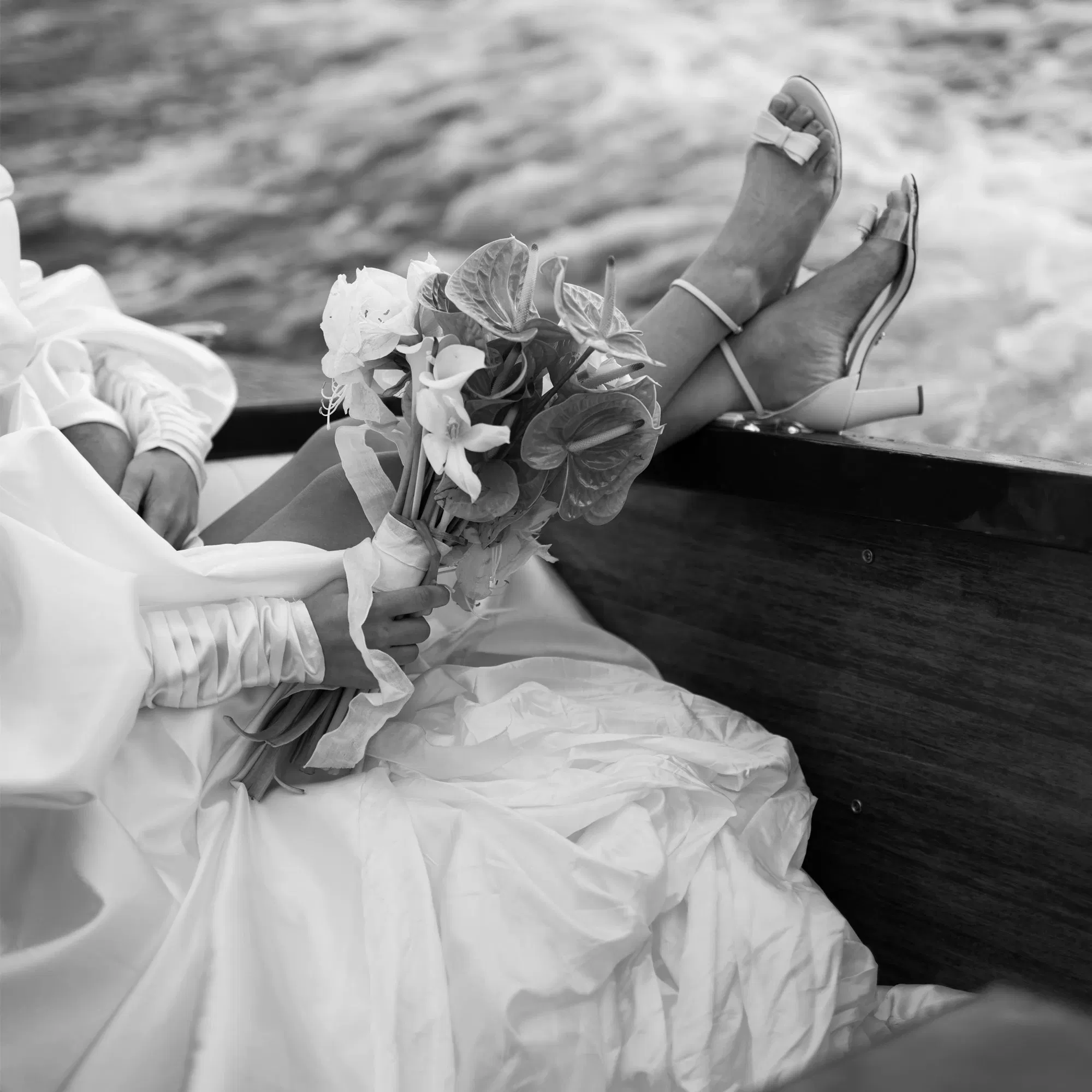Schwarz-Weiß-Fotografie einer Braut in einem Boot, die einen eleganten Blumenstrauß hält und ihre Schuhe mit Schleifen-Detail zeigt.