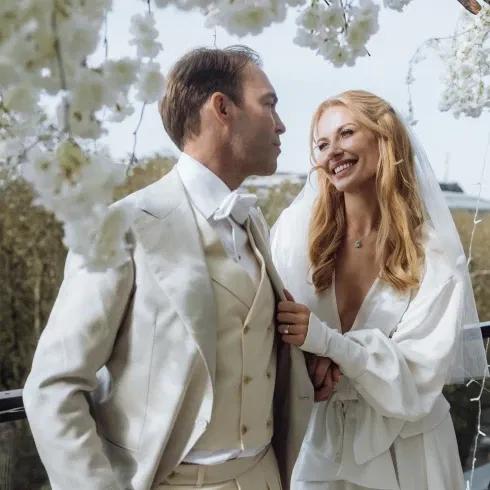 Das Brautpaar in harmonischen, hellen Hochzeitsoutfits teilt einen intimen Augenblick auf einem Balkon mit blühender Dekoration.