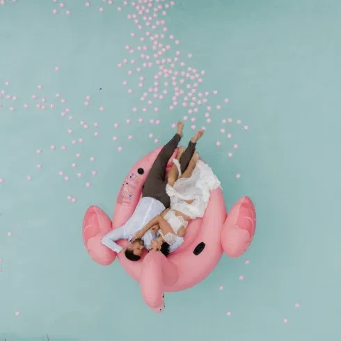 Brautpaar liegt entspannt auf einem pinken Flamingo-Schwimmtier in einem Pool, umgeben von rosa Wasserbällen.