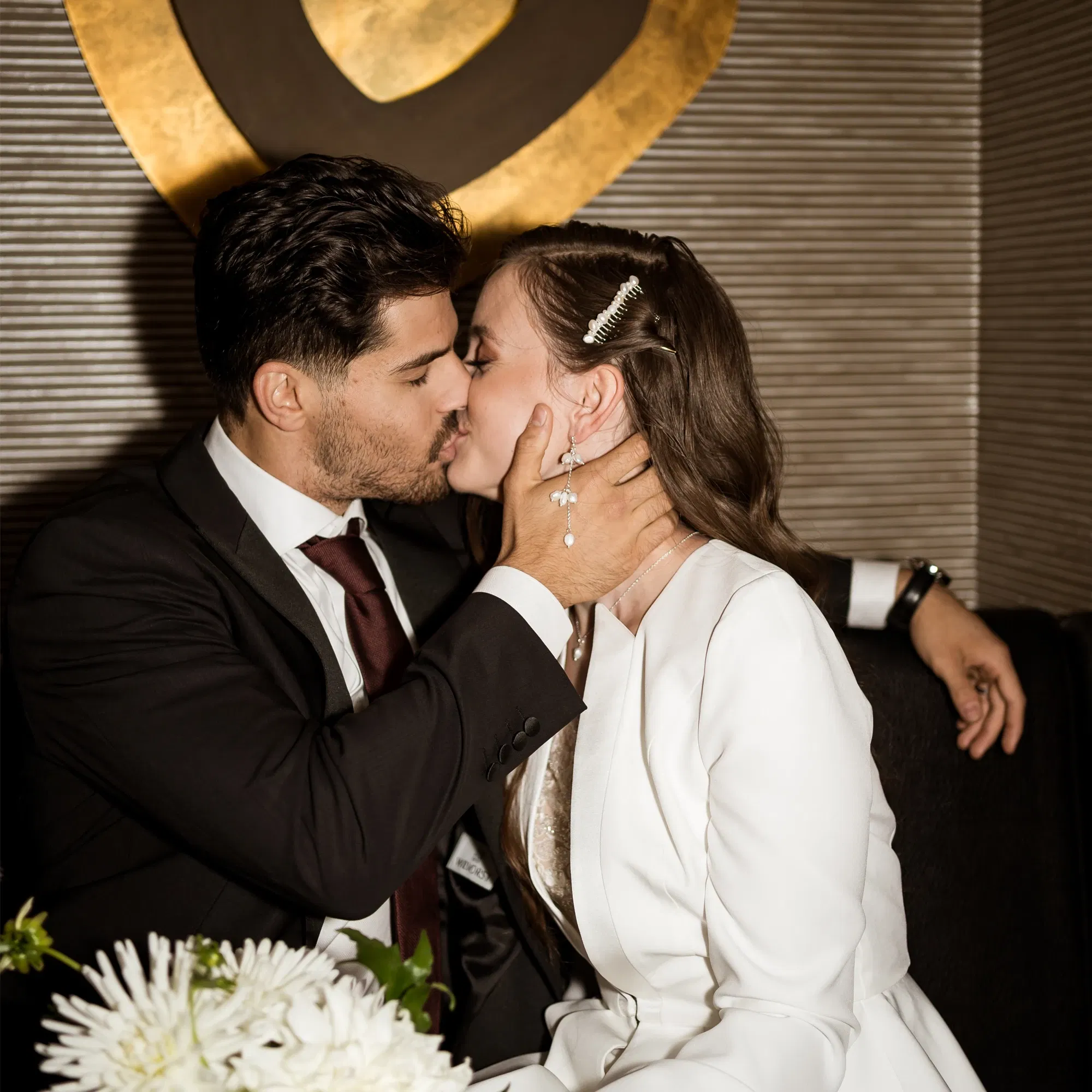 Ein Paar in eleganter Abendgarderobe, das sich innig küsst. Die Braut trägt eine stilvolle weiße Jacke mit Haarspange und auffälligen Ohrringen. Romantischer Moment in urbanem Ambiente.