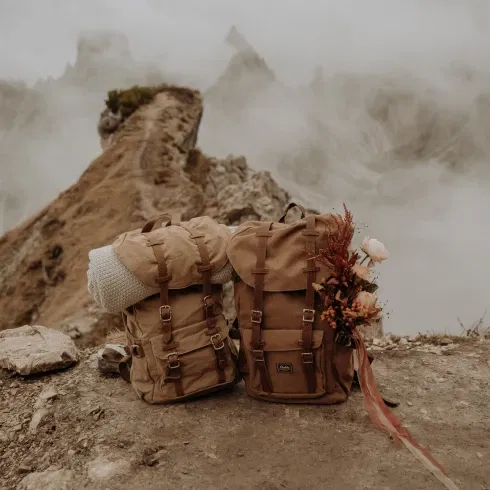 Zwei beige Wanderrucksäcke auf einem felsigen Bergpfad mit einer nebligen Berglandschaft im Hintergrund, einer der Rucksäcke ist mit einem Blumenstrauß dekoriert.