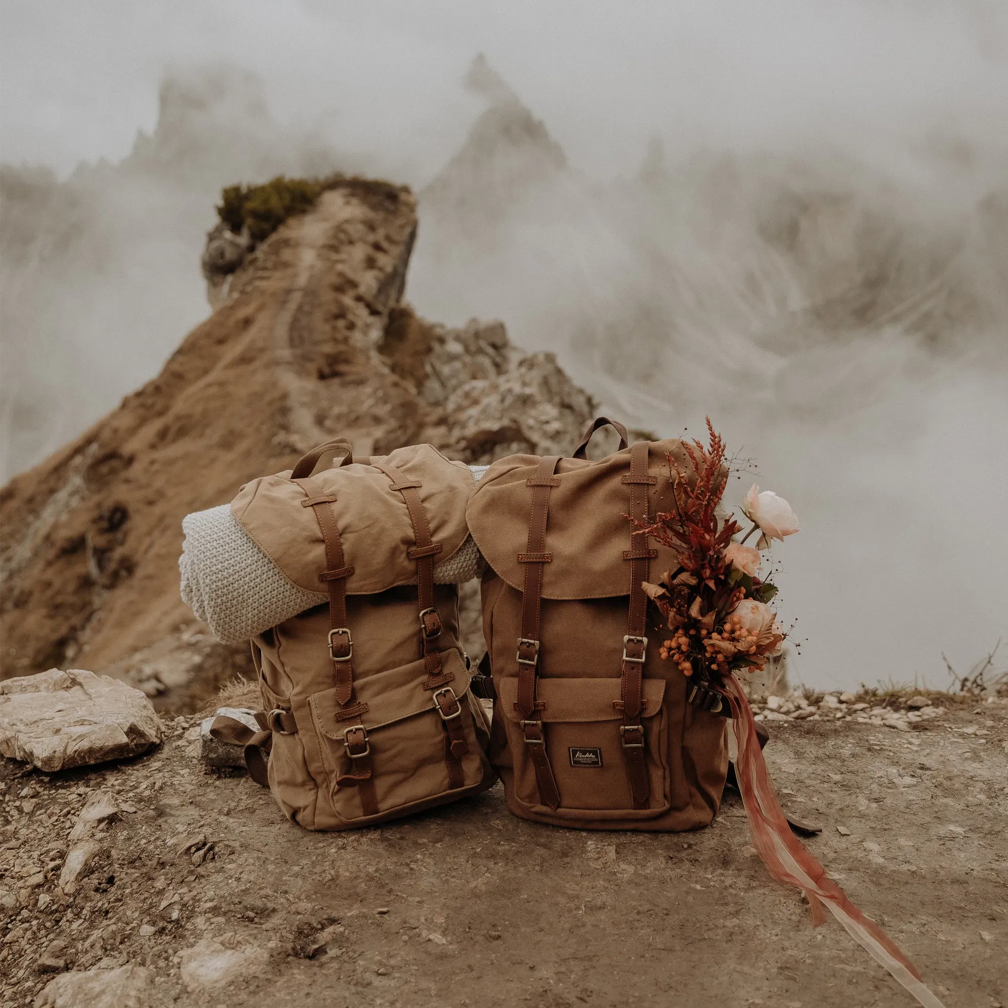 Zwei beige Wanderrucksäcke auf einem felsigen Bergpfad mit einer nebligen Berglandschaft im Hintergrund, einer der Rucksäcke ist mit einem Blumenstrauß dekoriert.