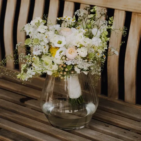 Ein üppiger Hochzeitsstrauß mit weißen und pastellfarbenen Blüten steht in einer klaren Vase auf einer hölzernen Bank, sanft beleuchtet.