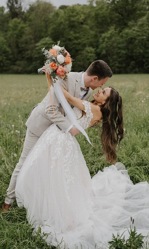 Braut und Bräutigam in einer romantischen Pose auf einer grünen Wiese, Braut in einem Spitzenkleid mit langem Schleier.