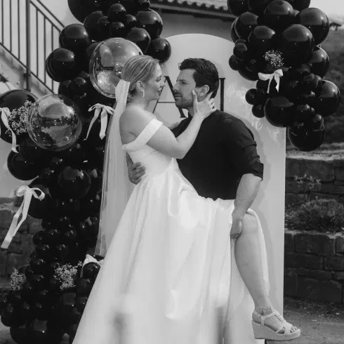 Ein Brautpaar in Schwarz-Weiß, das sich innig ansieht. Die Braut trägt ein trägerloses weißes Kleid und einen Schleier, während der Bräutigam in einem schwarzen Hemd sie hält. Im Hintergrund befindet sich eine Dekoration aus schwarzen Ballons und ein weißer Bogen.