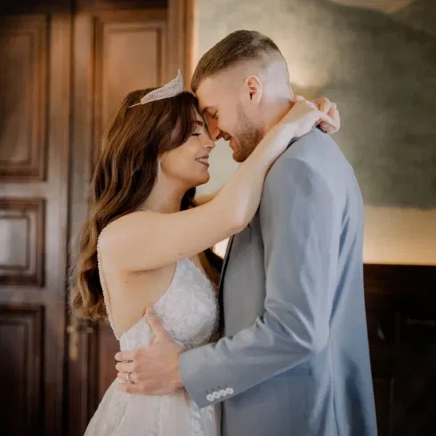 Braut und Bräutigam in einem intimen Moment, lächelnd und umarmt. Die Braut trägt ein funkelndes Diadem und ein elegantes Spitzenkleid, während der Bräutigam einen hellen Anzug trägt.