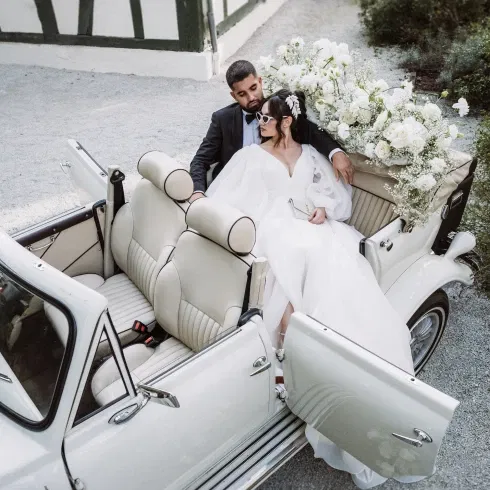 Brautpaar in einem cremefarbenen Oldtimer, die Braut in einem eleganten Kleid mit Sonnenbrille, umgeben von weißen Blumenarrangements im Cabrio.