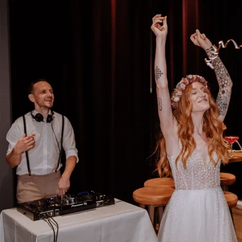 Eine Braut mit Blumenkranz und einem transparenten, bestickten Brautkleid tanzt fröhlich neben einem DJ, der lächelnd auflegt.