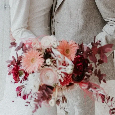 Nahaufnahme eines Brautstraußes mit rosa Gerbera, cremefarbenen Ranunkeln und roten Akzenten, gehalten von der Braut in einem eleganten Kleid.