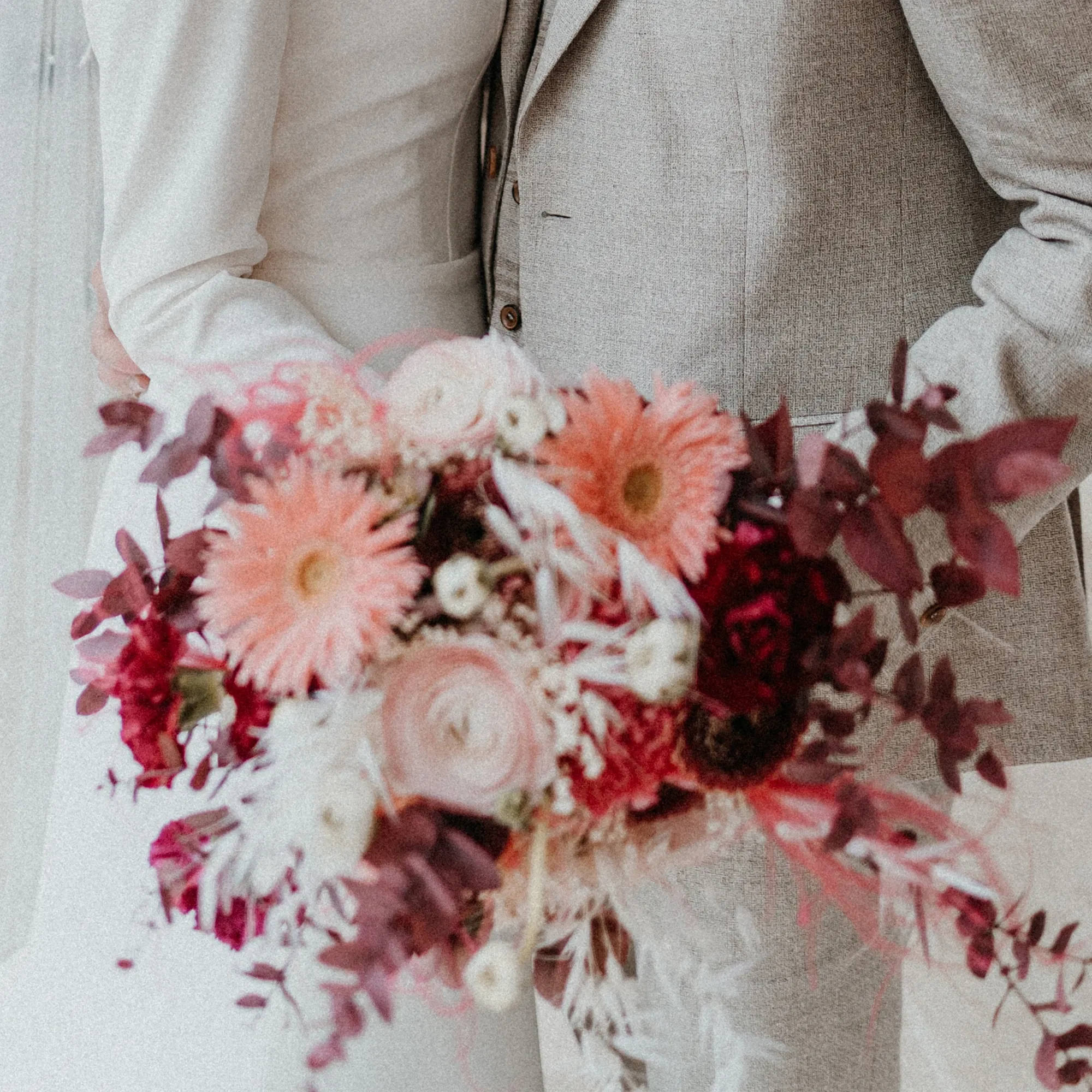 Nahaufnahme eines Brautstraußes mit rosa Gerbera, cremefarbenen Ranunkeln und roten Akzenten, gehalten von der Braut in einem eleganten Kleid.